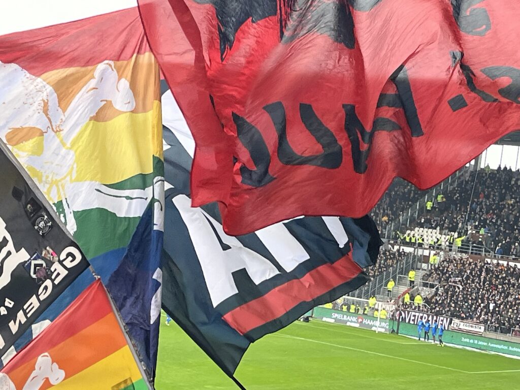 Fahnenmeer bei einem Heimpiel des FC St. Pauli, zu erkennen eine Regenbogen-Fahne mit Totenkopf und eine rote Fahne mit FC St. Pauli - Aufdruck