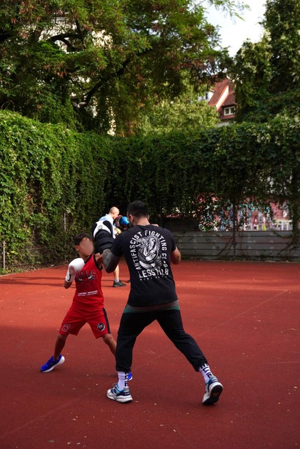 Sportplatz. im Vordergrund ein Trainer mit Pratzen, von hinten zu erkennen, trainierend mit einem Kind. Auf einem Sportplatz mit rotem Belag