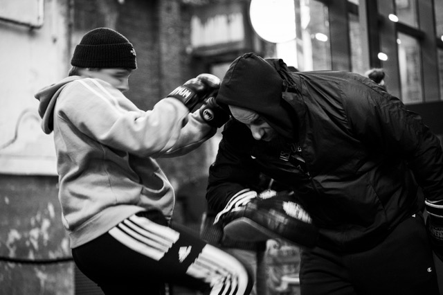 Schwarz-Weiß-Bild, ein Kind im Trainingsanzug, mit Hoodie, Mütze und Boxhandschuh führt einen Kick mit dem Bein aus, der von einem Trainer, ebenfalls im Hoodie, abgewehrt wird