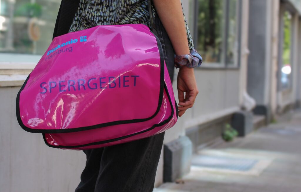 Ein Mensch steht an der Straße, umgehängt eine Messenger-Tasche, beschriftet mit "Diakonie Hamburg, Sperrgebiet"