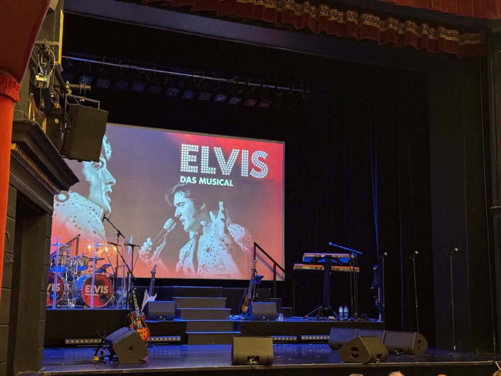 Blick auf eine Theaterbühne vor Showbeginn mit Musikinstrumenten und einer großen Leinwand, auf der der Schriftzug „ELVIS DAS MUSICAL“ und ein Portrait von Elvis Presley zu sehen sind.