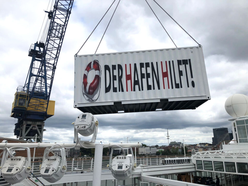 Ein Container am Kran, der Container per Bildbearbeitung beschriftet mit "Der Hafen hilft"