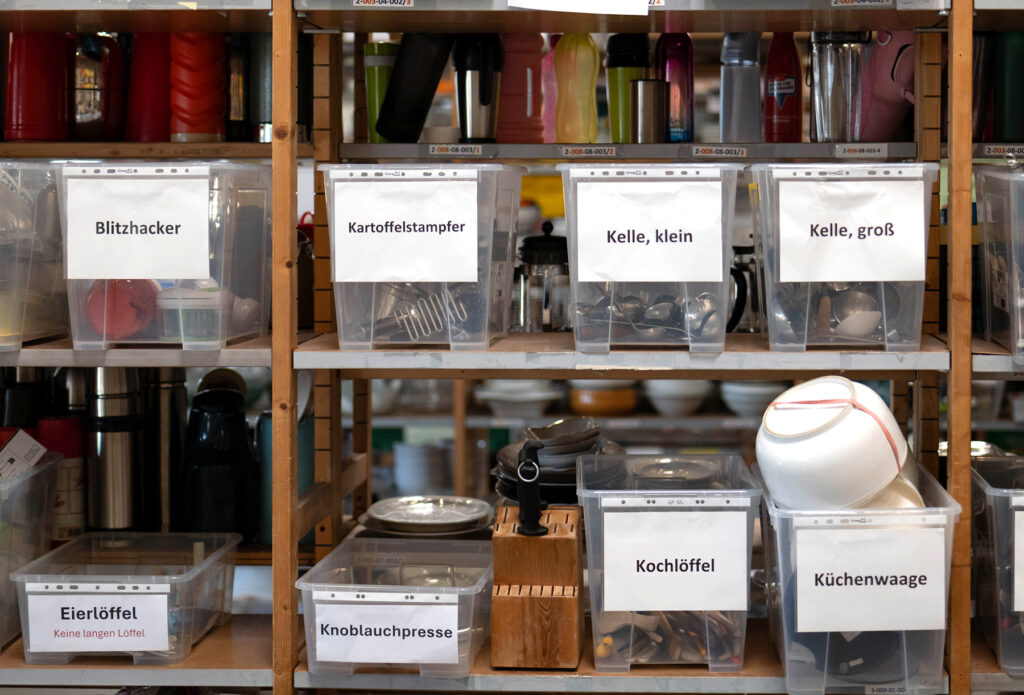 Regale, in denen in volle Plastikbehälter aufgereiht sind. Diese Behälter sind beschriftet mit "Blitzhacker", "Kartoffelstampfer", "Kelle, klein", "Kelle, groß", "Eierlöffel", "Knoblauchpresse", "Kochlöffel" und "Küchenwaage"