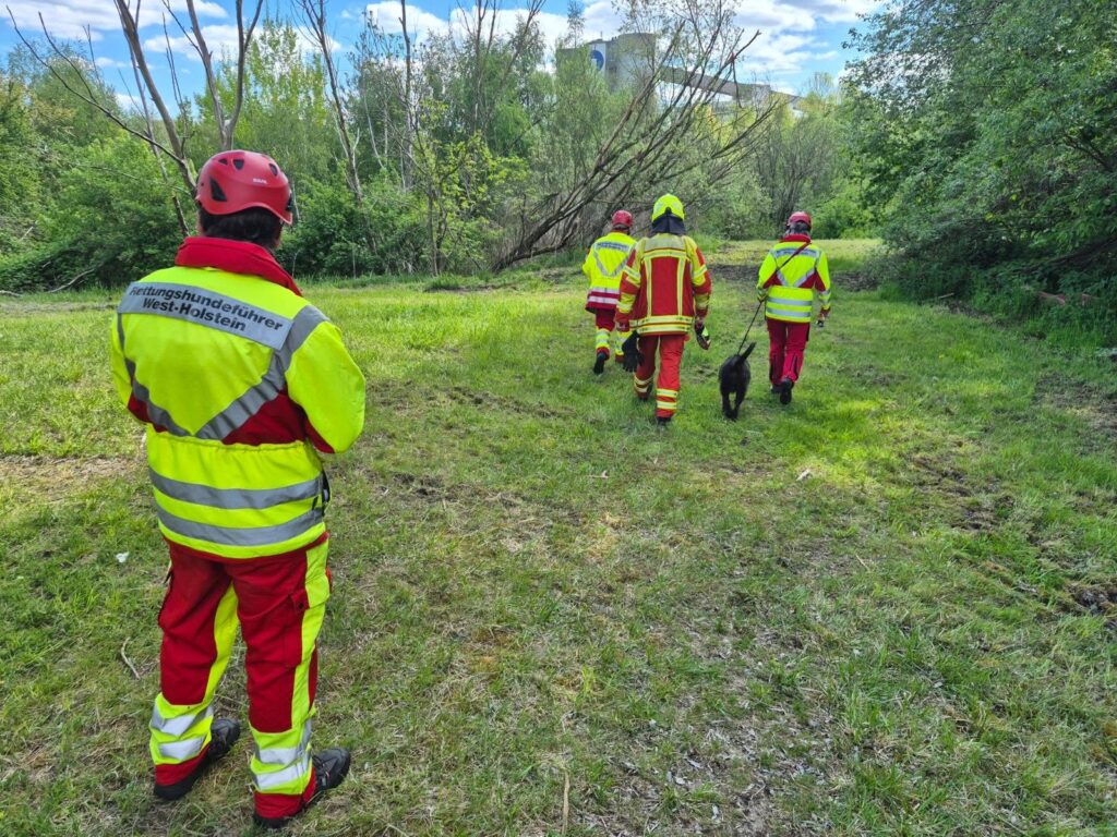 4 Personen von hinten zu erkennen in Jacken mit Signalfarben und Beschriftung "Rettungshundeführer West-Holstein"