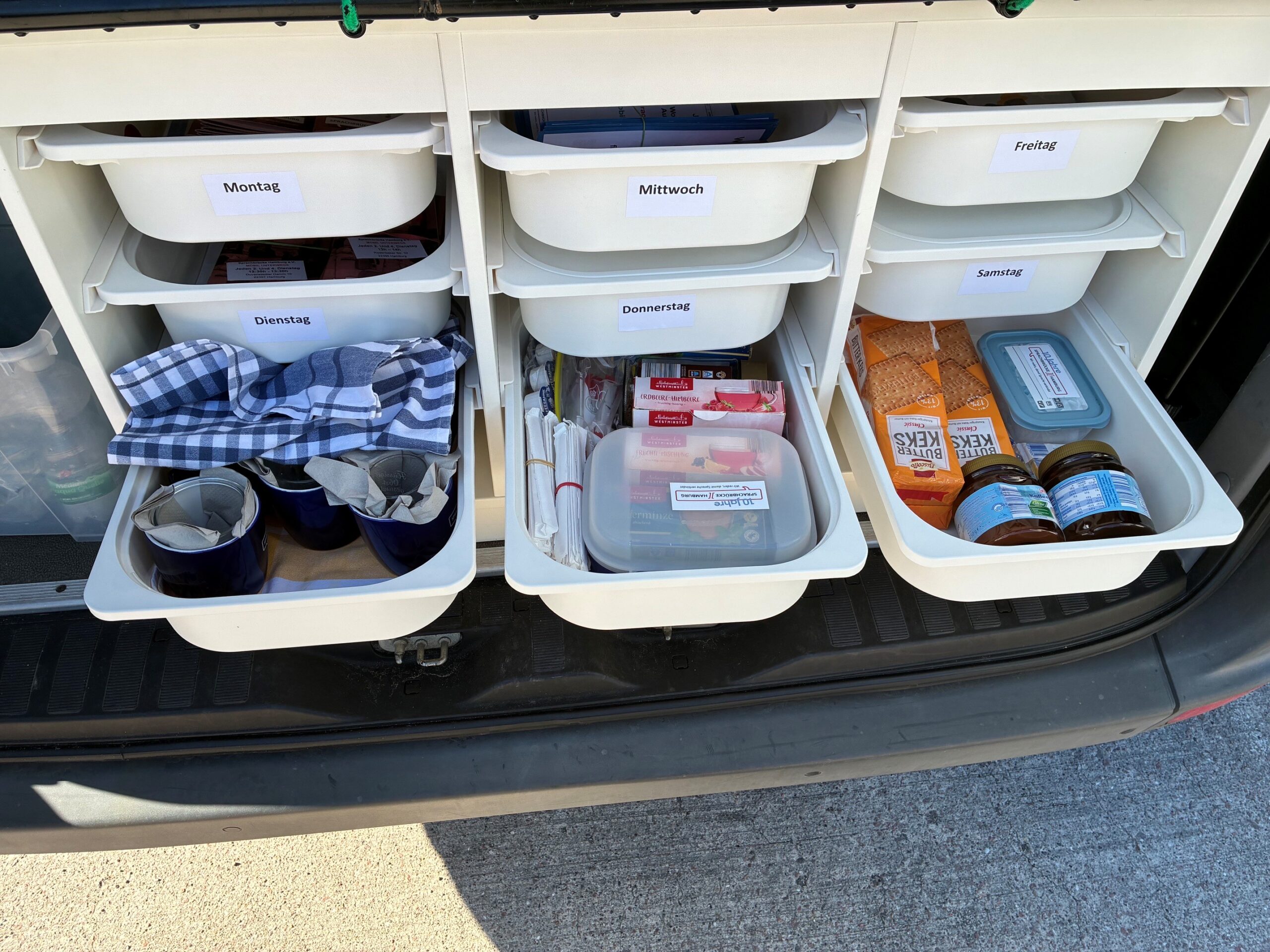 Es sind die Ablagefächer im Sprachbrücken-Mobil zu erkennen. Geschlossene Schubladen beschriftet mit "Montag" bis "Samstag" und geöffnete Schubladen mit Kaffeetassen, Keksen und Kaffee-Zubehör