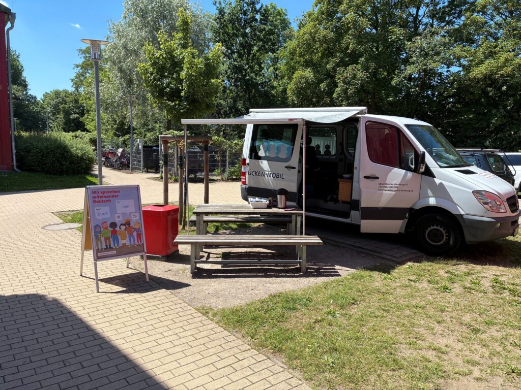 Sprachbrücken-Mobil mit ausgefahrener Markise und geöffneter Seitentür, davor eine Sitzgarnitur und Aufsteller "Wir sprechen miteinander Deutsch"