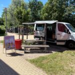 Sprachbrücken-Mobil mit ausgefahrener Markise und geöffneter Seitentür, davor eine Sitzgarnitur und Aufsteller "Wir sprechen miteinander Deutsch"